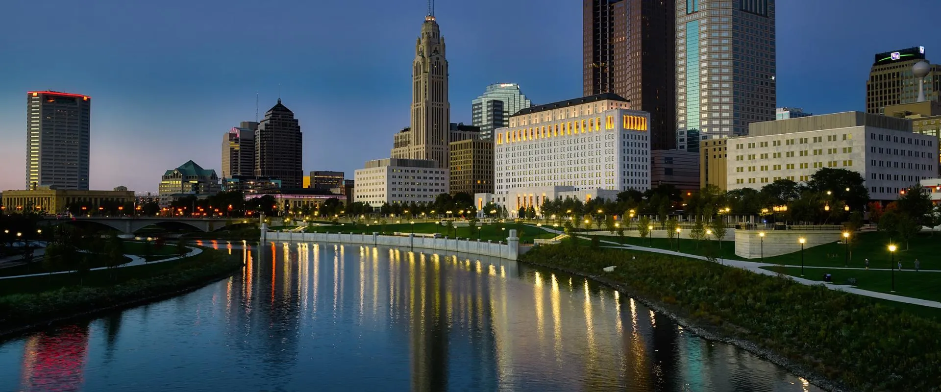 A river flows through a cityscape at dusk, with buildings silhouetted against a colorful evening sky.